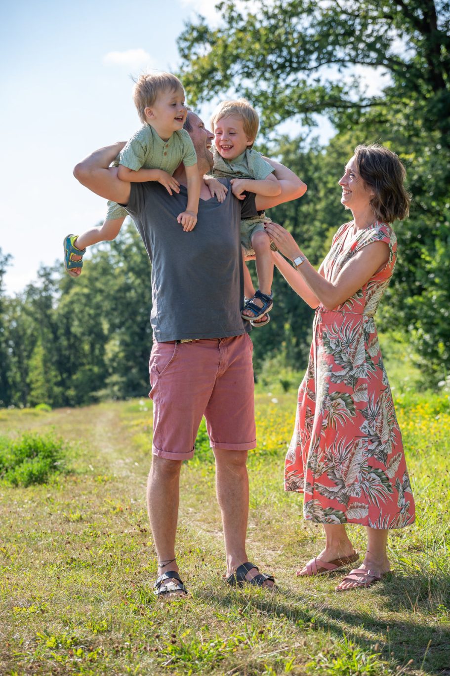 Familie in der Natur: Vater trägt zwei fröhliche Kinder, Mutter steht daneben.