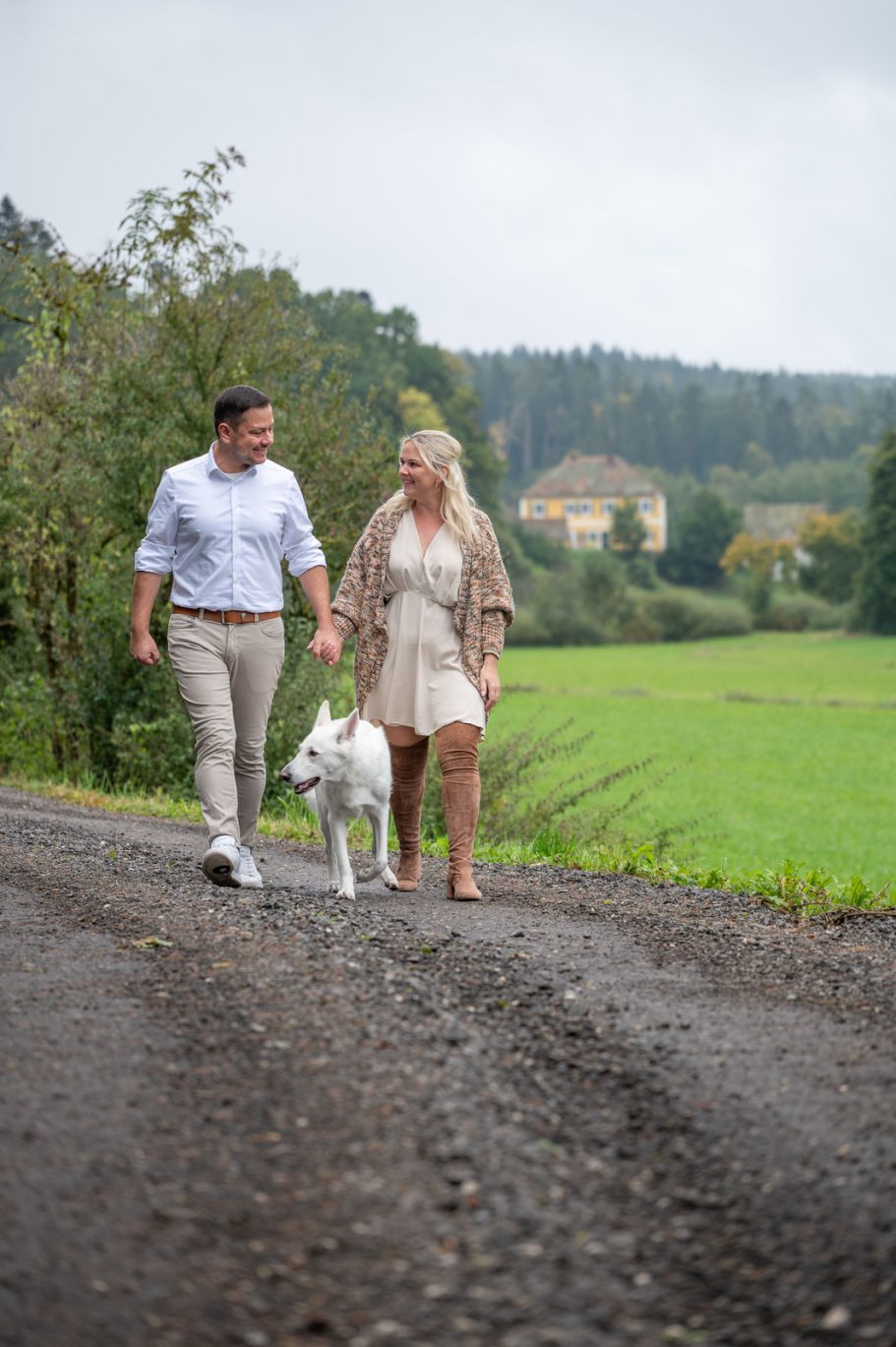 Paar mit Hund spaziert auf einem Weg durch eine grüne Landschaft.