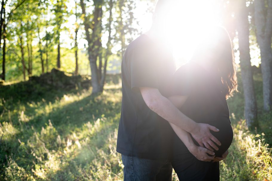 Babybauchshooting - Mann fasst Frau an den Babybauch, die Sonne scheint in die Kamera