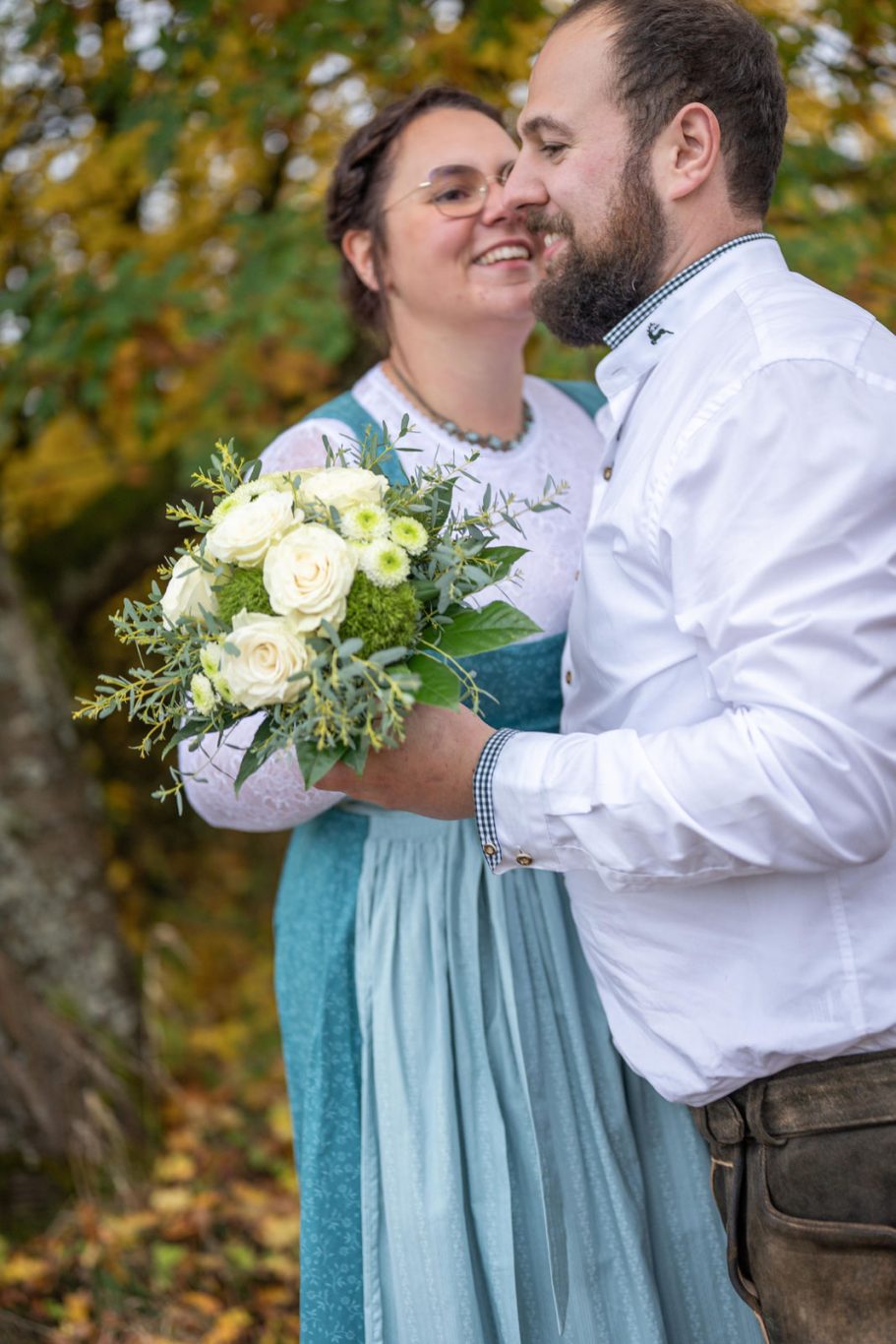 Paar umarmt sich lächelnd, Braut hält einen Strauß mit weißen Blumen. Herbstliche Umgebung.