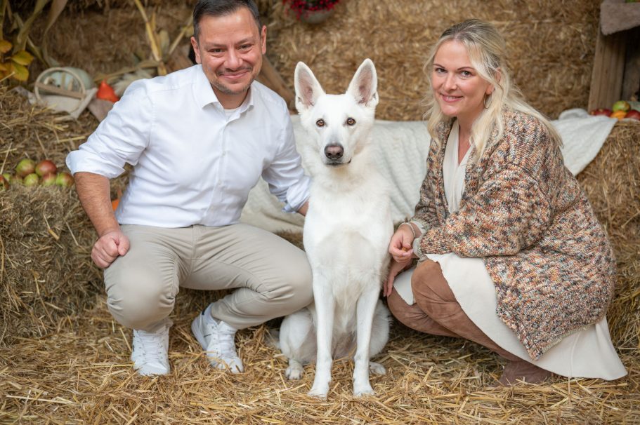 Paar mit einem weißen Hund posiert vor einer strohgedeckten Kulisse.