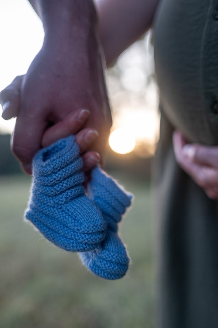 BABYBAUCH Hände halten blaue Baby-Schuhe vor einer schwangeren Person im Sonnenuntergang.