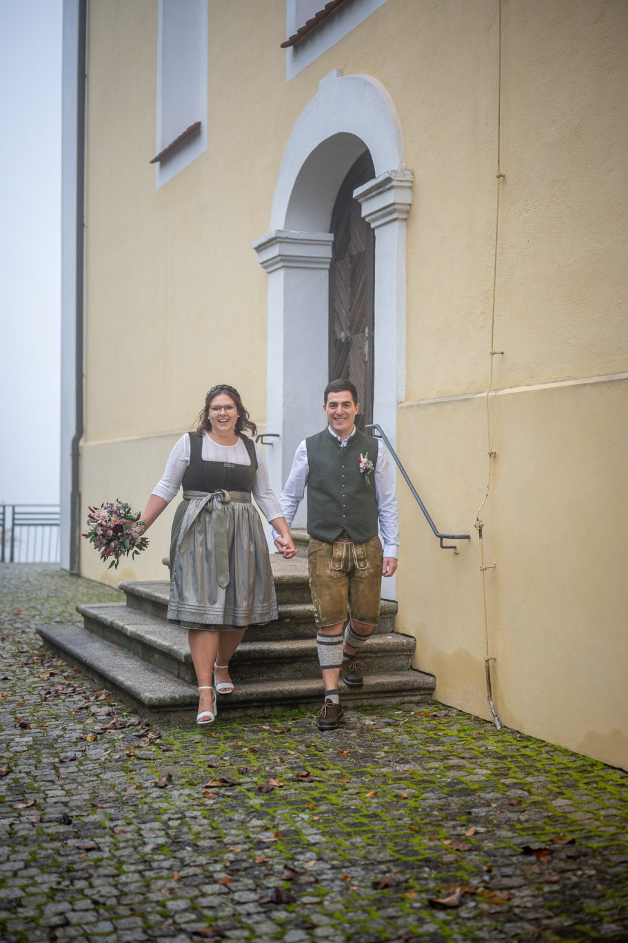 Brautpaar in traditioneller Tracht, Hand in Hand vor einem historischen Gebäude.