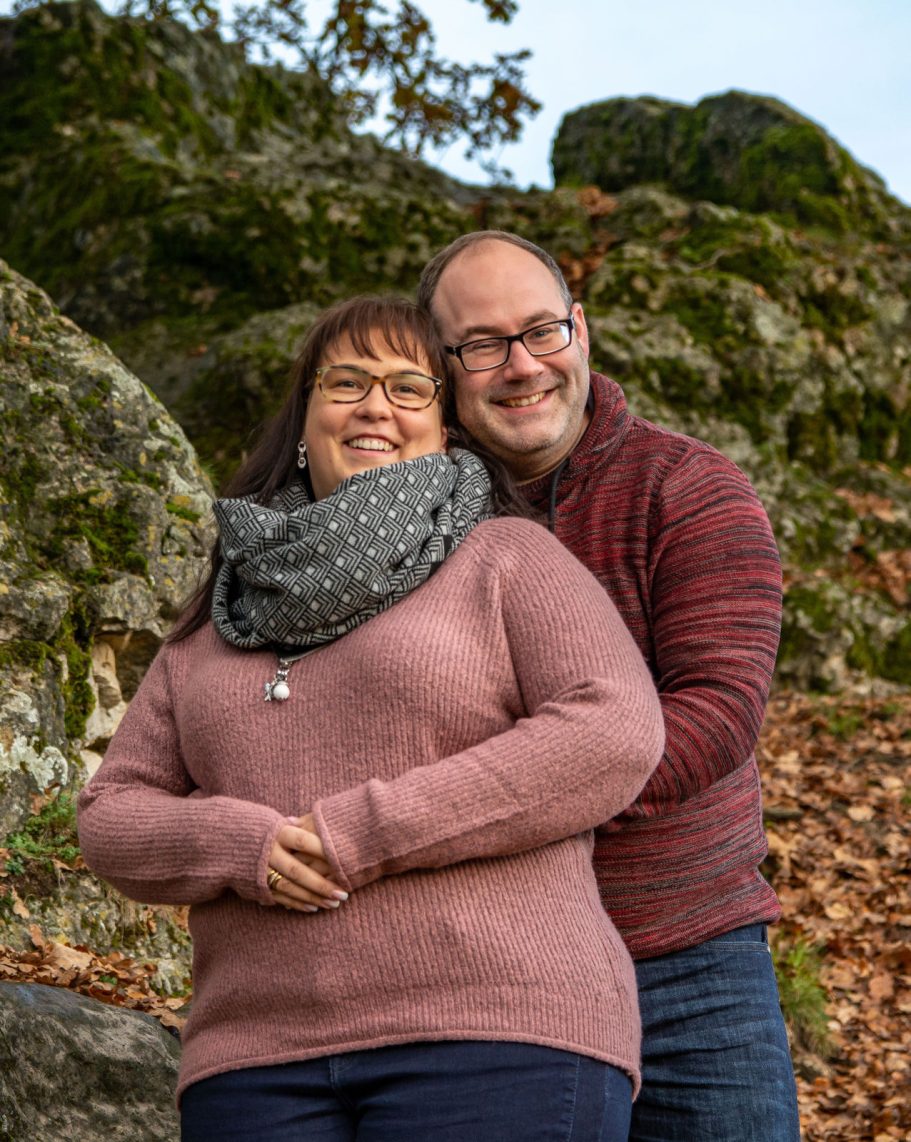 Ein Paar steht umarmt vor einer felsigen Kulisse im Freien, umgeben von herbstlichen Blättern.