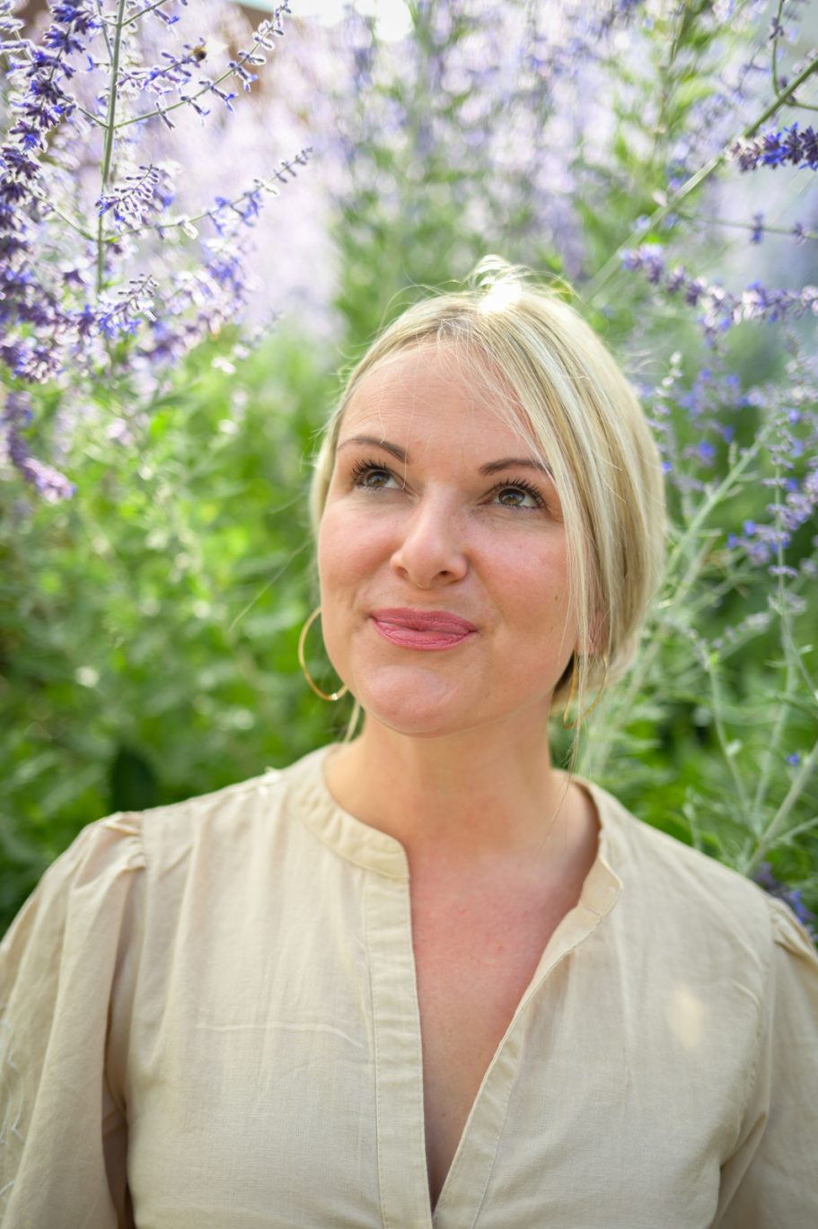 Frau mit blonden Haaren, lächelnd vor lila blühenden Pflanzen.