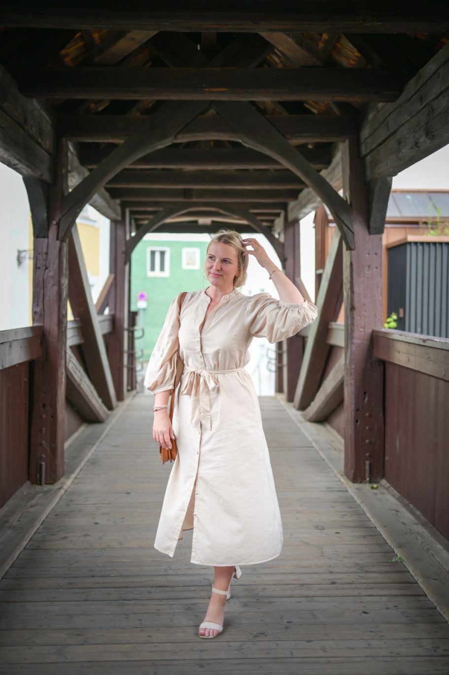 Frau in einem beigen Kleid geht über eine Holzbrücke.