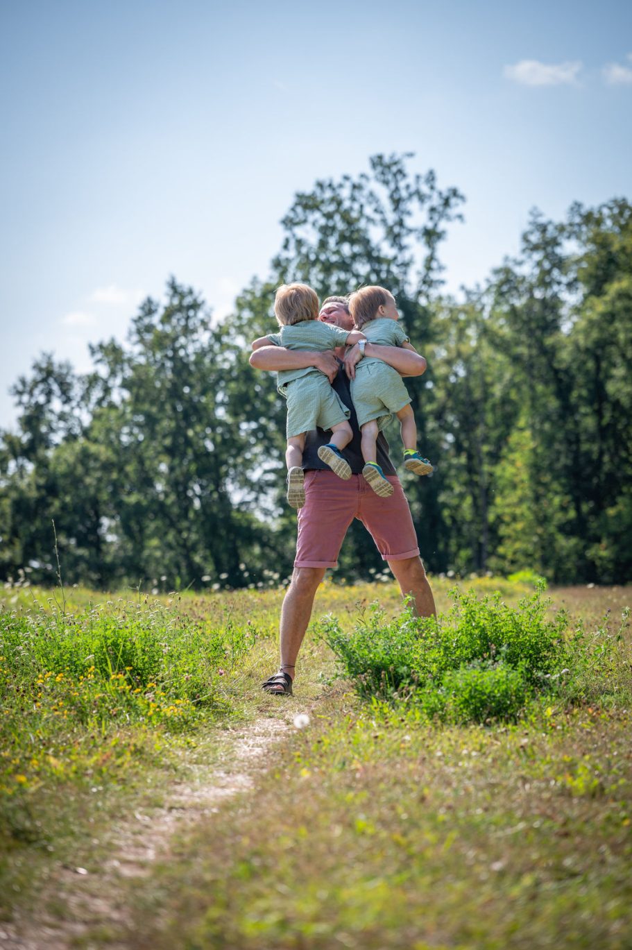 Mann trägt zwei Kleinkinder auf dem Rücken in einer grünen Wiese.