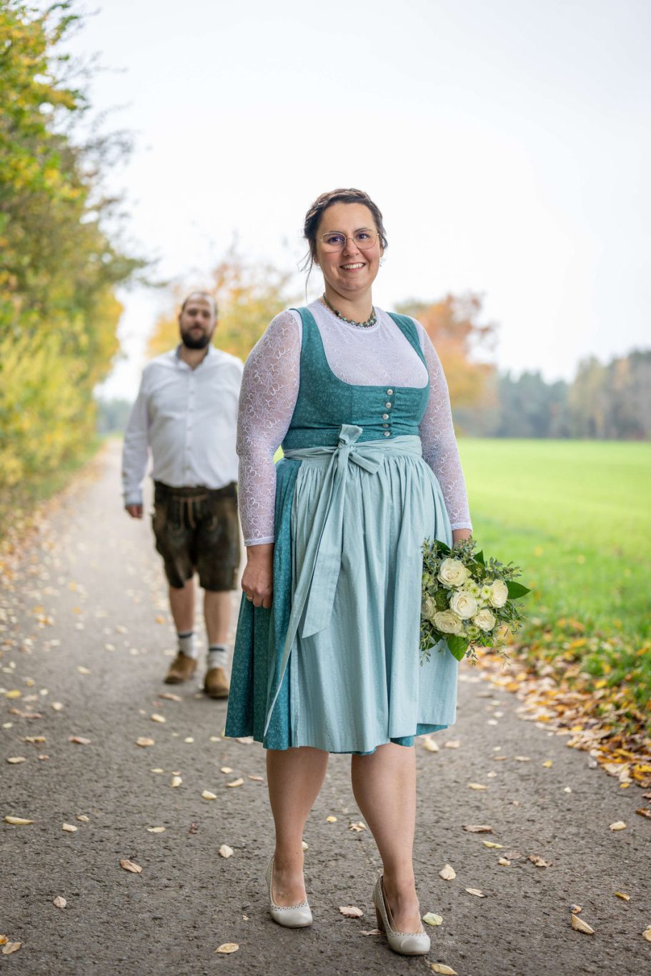 Braut in traditioneller Tracht mit einem Brautstrauß, der Bräutigam folgt im Hintergrund.