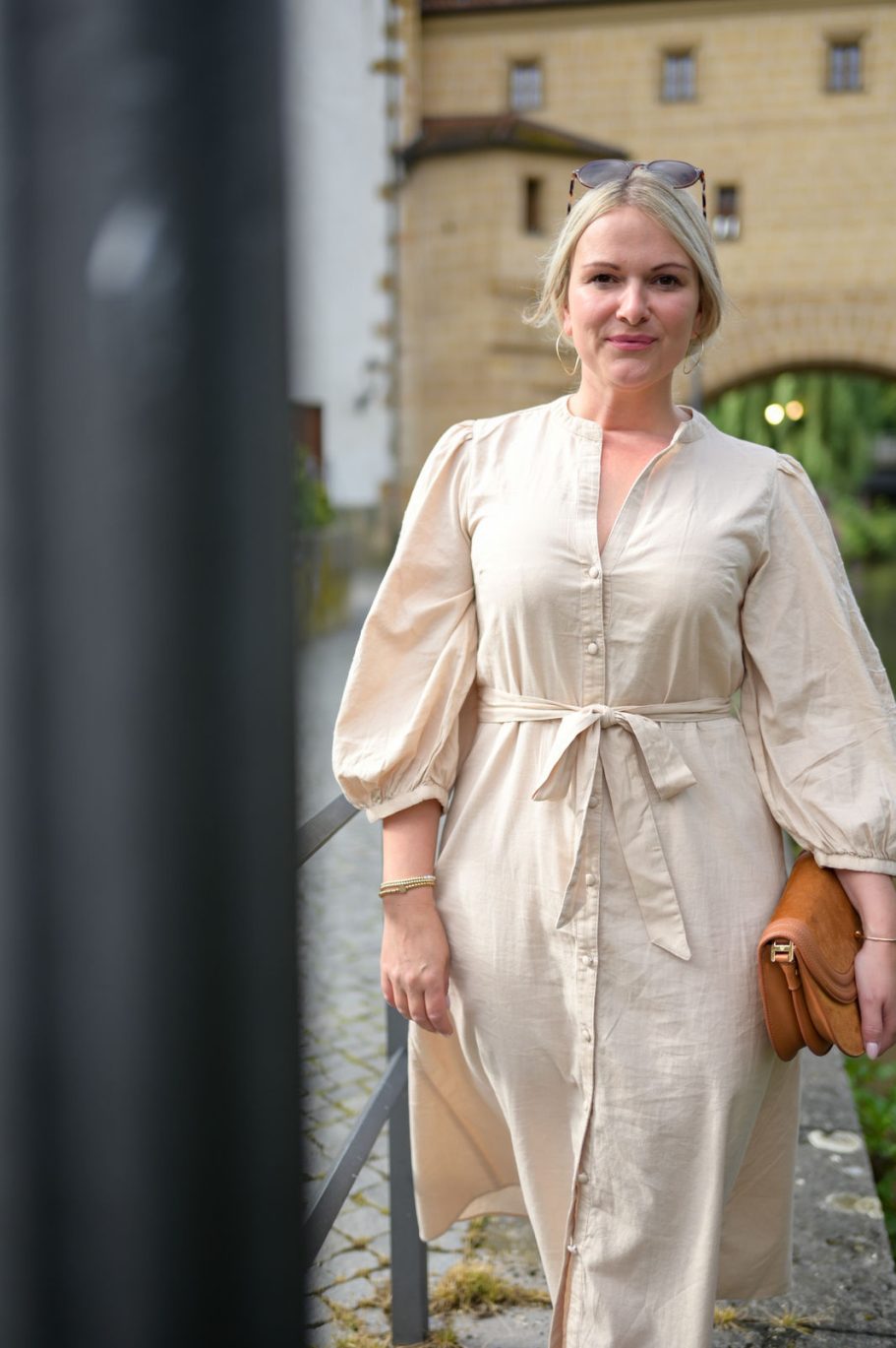 Frau in einem beigen, taillierten Kleid mit Armrüschen und einer braunen Tasche.