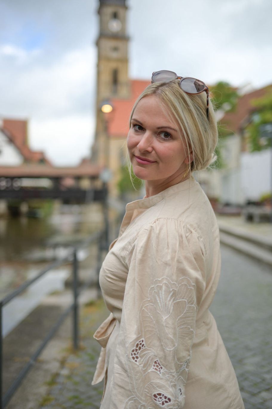 Frau mit blonden Haaren und Sonnenbrille vor einer Stadtansicht am Kanal.