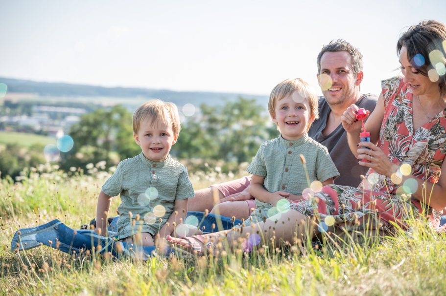 Familie sitzt im Gras und lächelt, mit zwei kleinen Kindern und bunten Spielsachen.