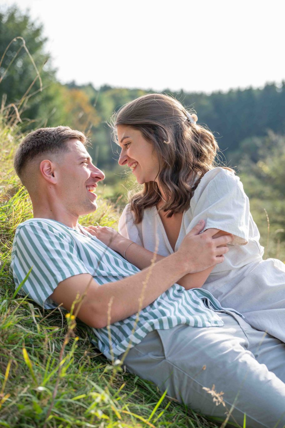 PAARE Junges Paar liegt im Gras, lächelt sich an, umgeben von einer naturnahen Landschaft.