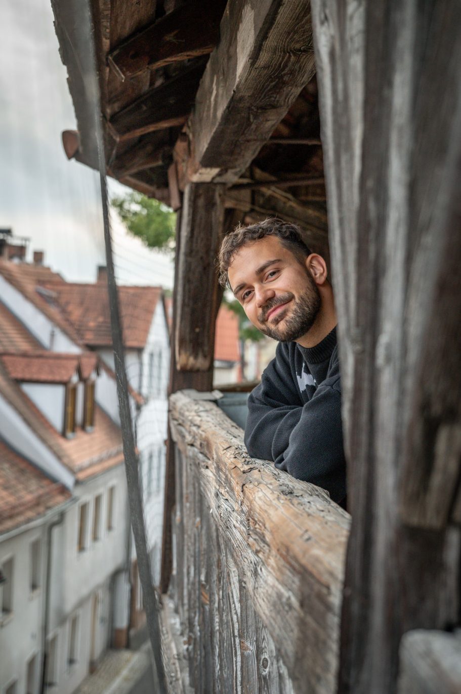 Mann lächelt von einem Balkon mit Blick auf historische Gebäude und eine Stadtlandschaft.