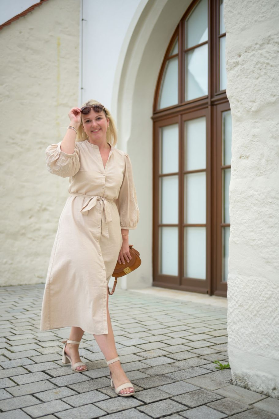 Frau in einem beigen Kleid geht lächelnd auf einem Pflasterweg.