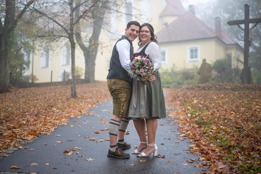 Brautpaar in traditioneller Tracht, umgeben von herbstlichem Laub und Nebel.