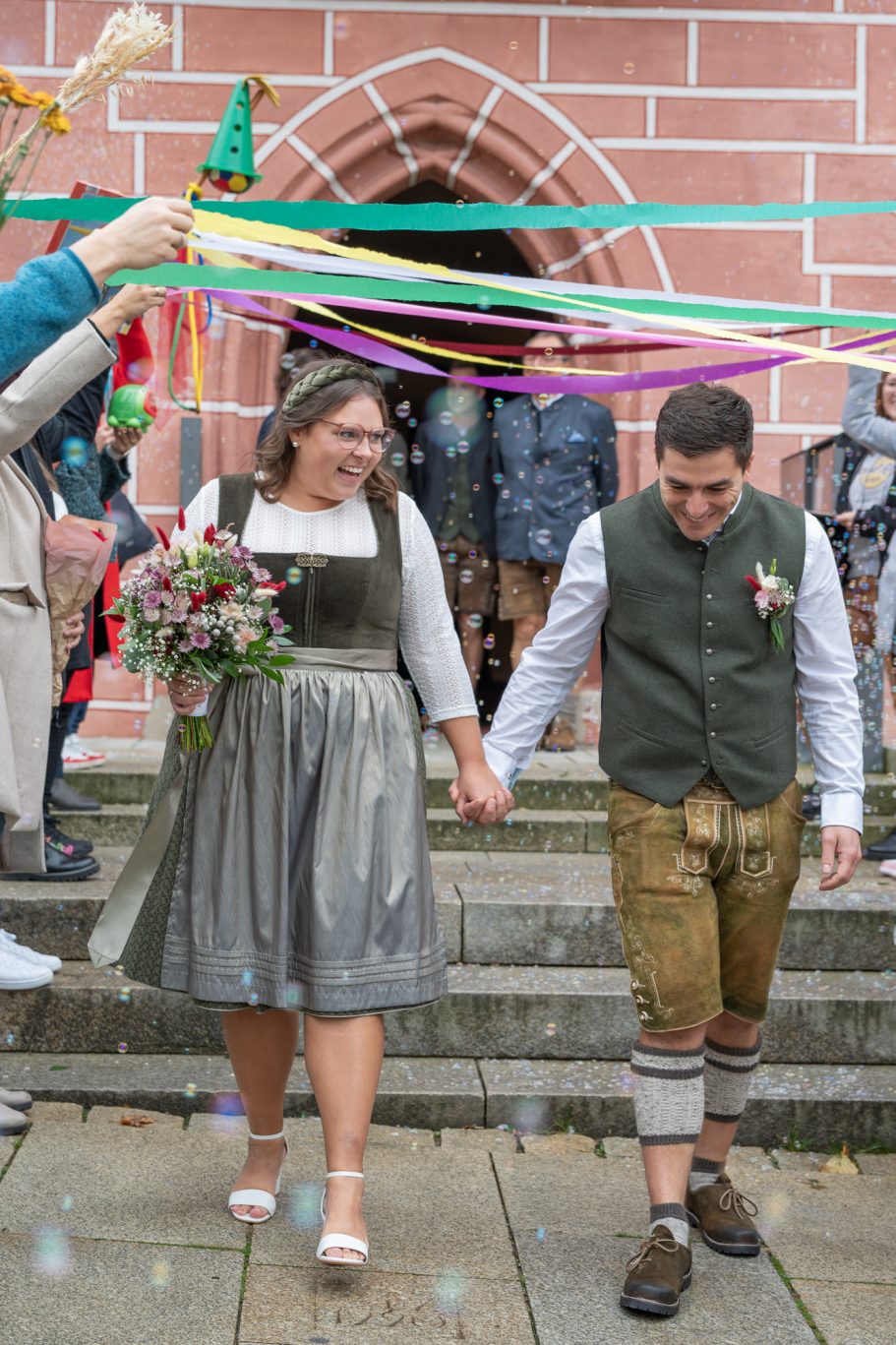 Ein Brautpaar in traditioneller Kleidung, Hand in Hand, umgeben von Feiernden und bunten Bändern.