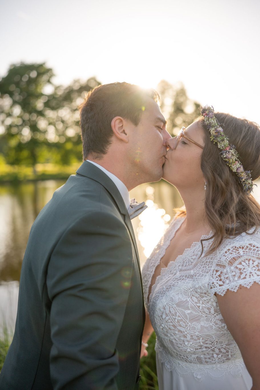 HOCHZEITEN Das Brautpaar küsst sich romantisch am Ufer eines Sees bei Sonnenuntergang.