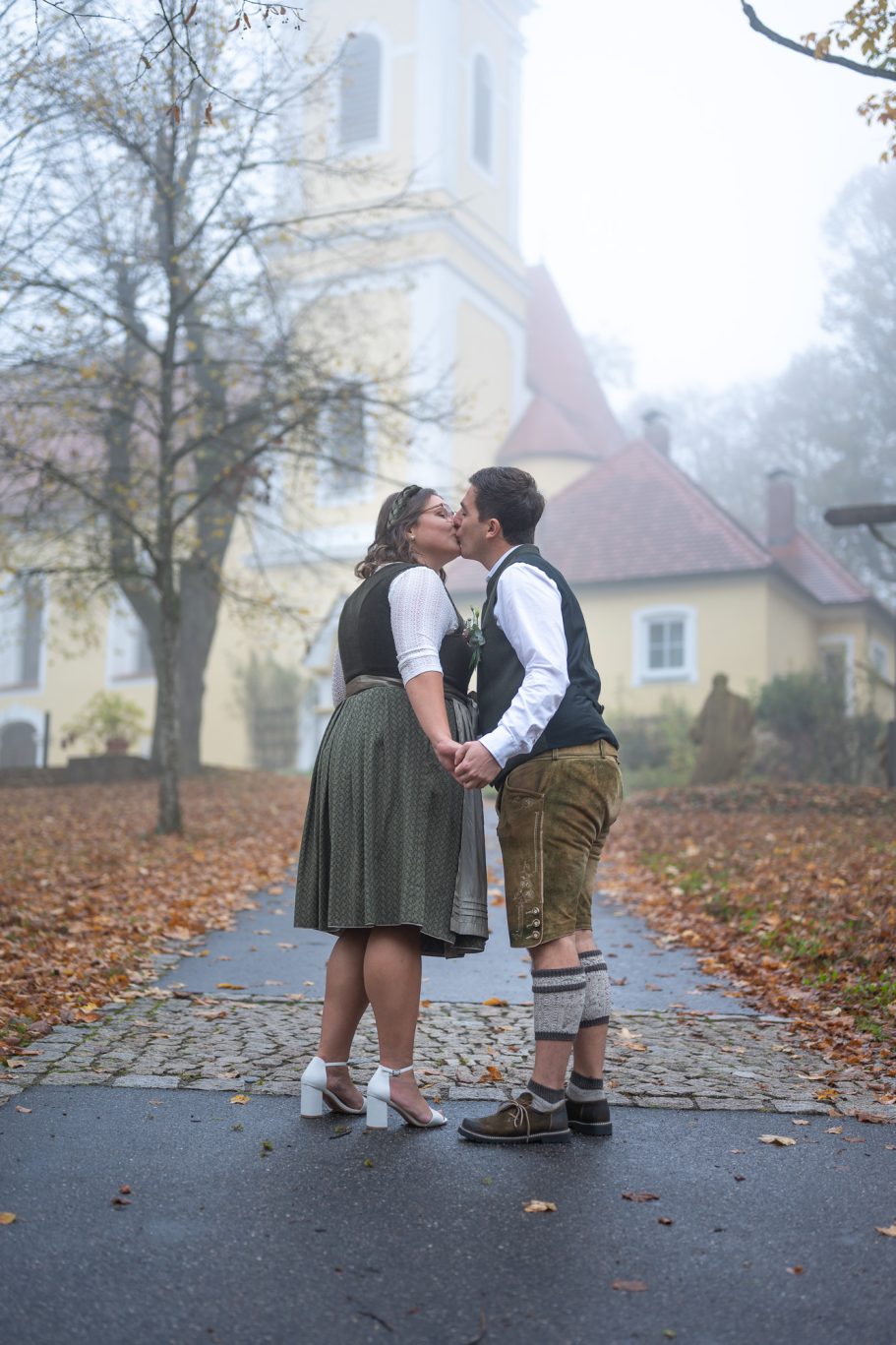 Paar in traditioneller Tracht küsst sich vor einer Kirche im Nebel.